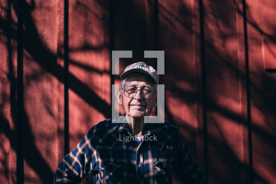An older man in a flannel shirt near a red barn wall.