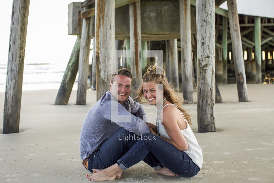 a couple sitting on a beach under a pier 