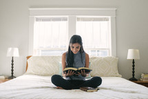 A smiling teen girl reading the Bible while sitting on her bed.