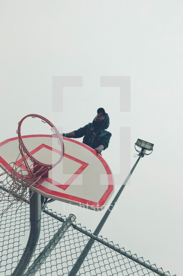 teen boy sitting on the top of a basketball goal 