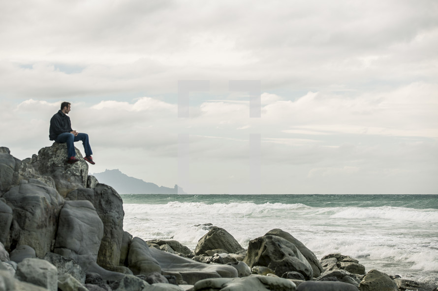 A man sitting atop huge boulders on a rocky shore.