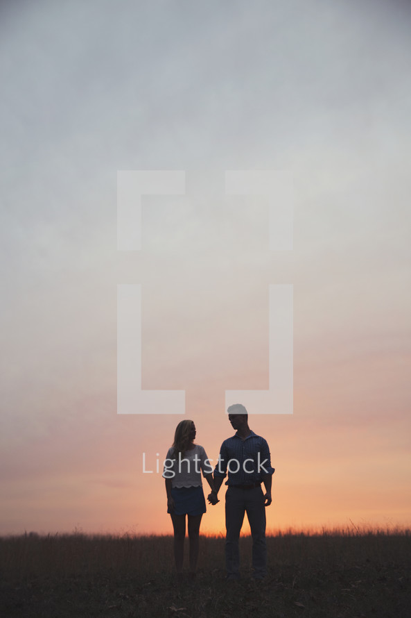 A man and woman holding hands in a field in front of a sunset.