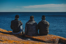 friends sitting on a rock on a shore looking out at the sea 