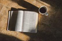 An open Bible lying on a tile floor and coffee mug 