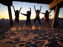 teen girls jumping in the sand under a pier on a beach 