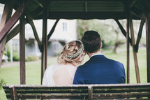 a bride and groom sitting on a bench outdoors with backs to the camera 
