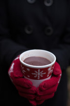 Hands wearing red mittens holding a cup of hot chocolate in a Christmas cup.