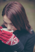 A young woman in red gloves drinks hot chocolate from a Christmas cup.