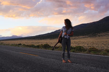 a young woman standing in the middle of a road holding a guitar 