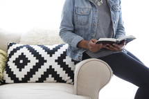 An African American woman sitting on the arm of a chair reading a Bible 