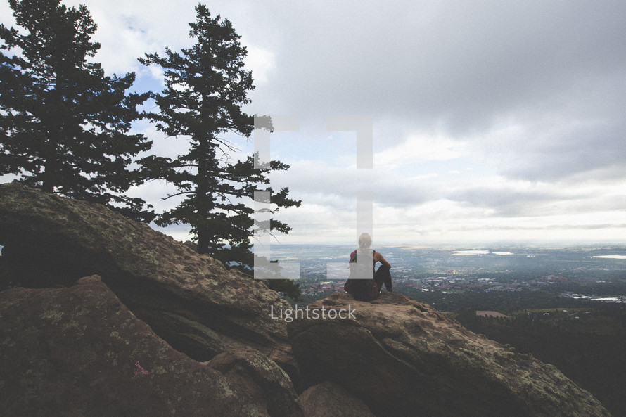 a woman sitting at the edge of a mountain cliff looking out at the valley below 