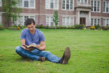 A man reads a Bible on a lawn outside an apartment building.