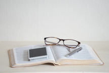 reading glasses, iPhone, and pen on the pages of a Bible 