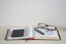 reading glasses, phone, and pen on the pages of a Bible 