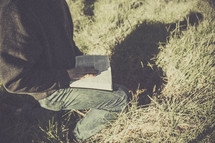  man sitting outdoors in tall grass reading a Bible  