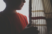 A boy stands near a window curtain.