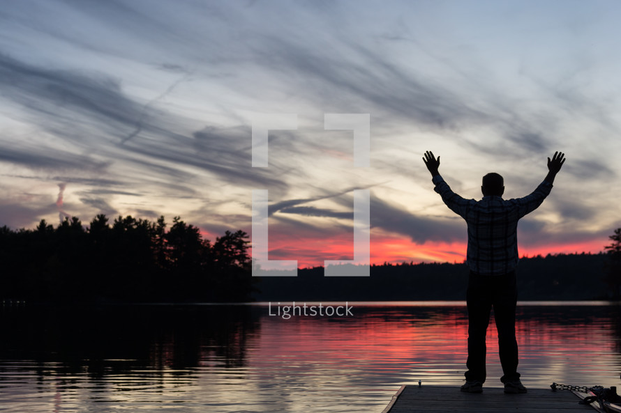 Silhouette of  a man with arms raised in praise at sunset over a lake.