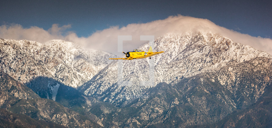 A yellow single engine airplane flying through snow covered mountains.