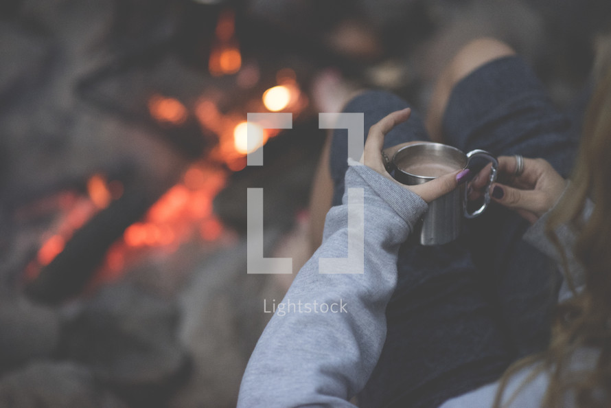 woman holding a mug of hot cocoa sitting by a campfire 