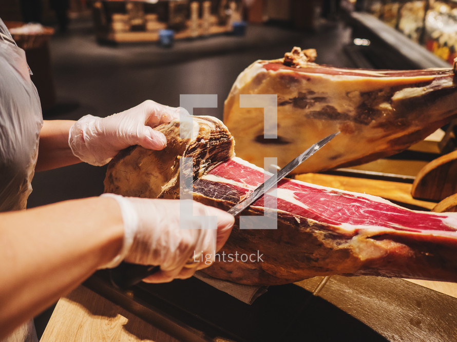 Hands of a woman in gloves in the market or store cut a slice of traditionally spanish jamon de bellota. Meat close up.