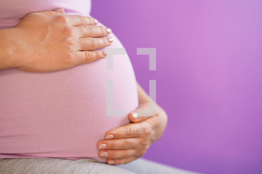 Pregnant woman holding her hands on tummy belly. Young girl expecting baby on pink bakcground. Maternity, motherhood, pregnancy concept.
