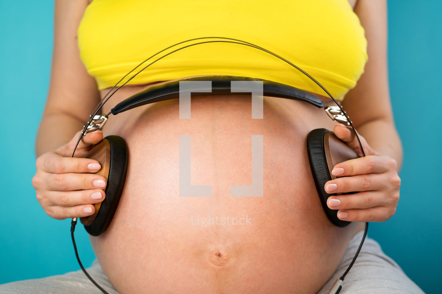 Pregnant woman holding earphones on her belly with baby, enjoying favorite music on blue background. Maternity, motherhood, pregnancy, love concept
