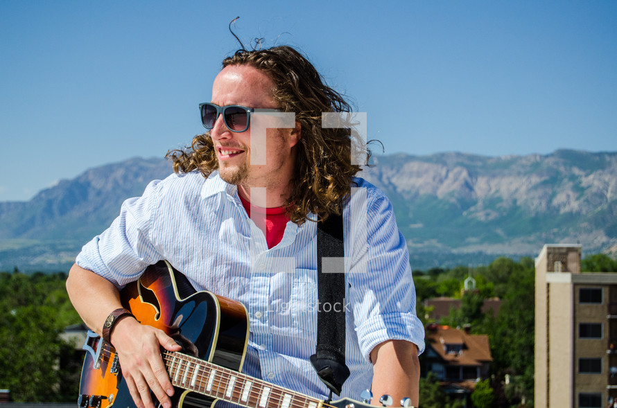 a man with a guitar and long hair in front of a mountain 