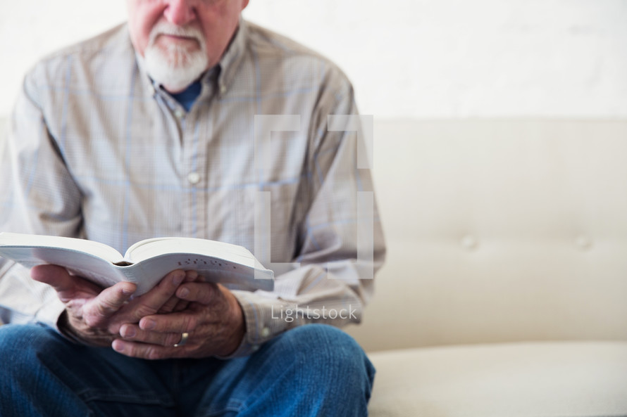 elderly man sitting on a couch reading a Bible 