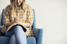 a woman sitting in a chair praying over an open Bible 