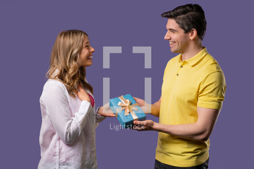 Young american couple on violet studio background. White man gives gift with bow to wife or girlfriend. Birthday, celebration, surprise, anniversary concept. High quality