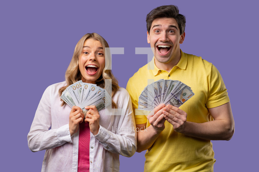 Amazed man and woman with USD currency. Young american student couple holding money - dollars banknotes on violet wall. Symbol of success, gain, salary, benefit, investments.