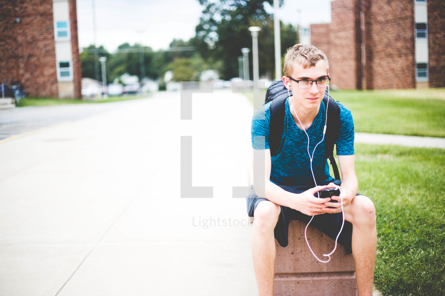a college student sitting with his backpack listening to earbuds 