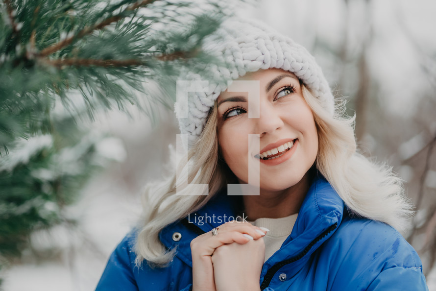 Portrait of young pretty woman in forest, winter season. Attractive kind blonde girl smiling, lady in white knitted hat. High quality
