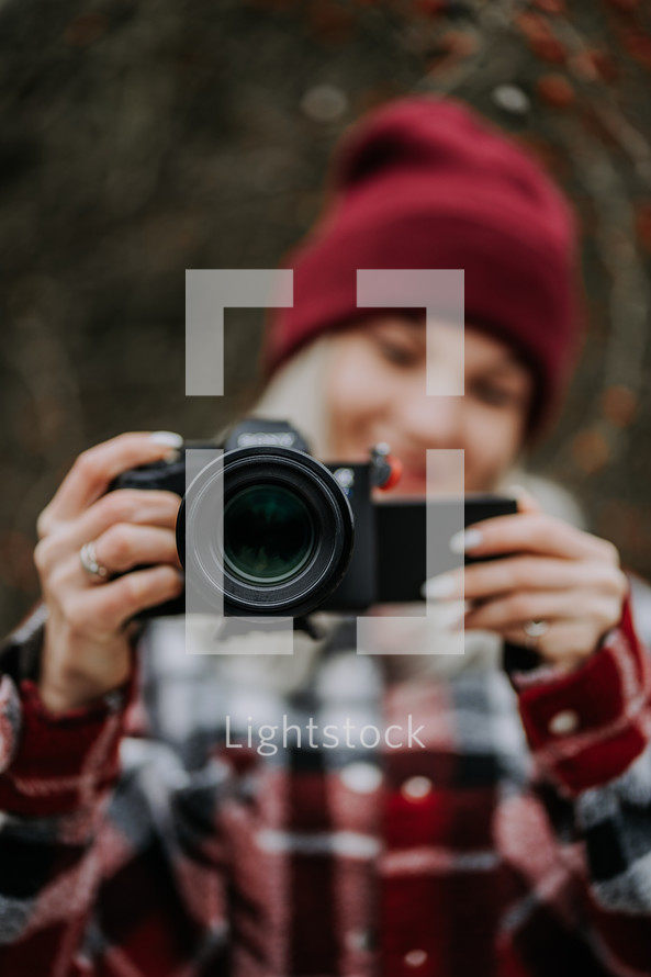 Portrait of young woman using camera in forest. Girl filming and smiling, lady in trendy shirt and red beanie hat. High quality