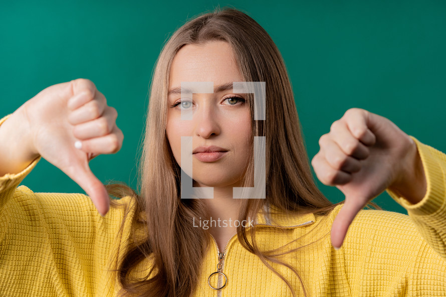 Portrait of unhappy european woman condemns with sign of dislike. Young millennial lady expressing discontent with showing thumbs-down gesture on blue studio background.