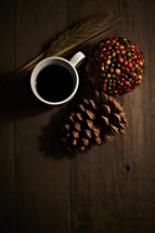 Cup of coffee sitting on wooden planks with pine cone, wheat stalk, and ball of berries.