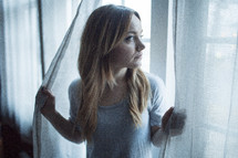 a young woman holding open curtains looking out a window 