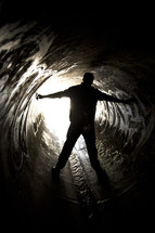 Silhouette of a man straddling water flowing through a sewer drain pipe with arms extended.