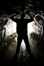 Man straddling water flowing through a sewer drain pipe with arms raised.