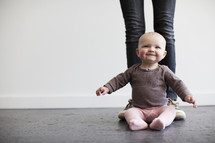 A baby sitting on the floor in front of a person's legs.