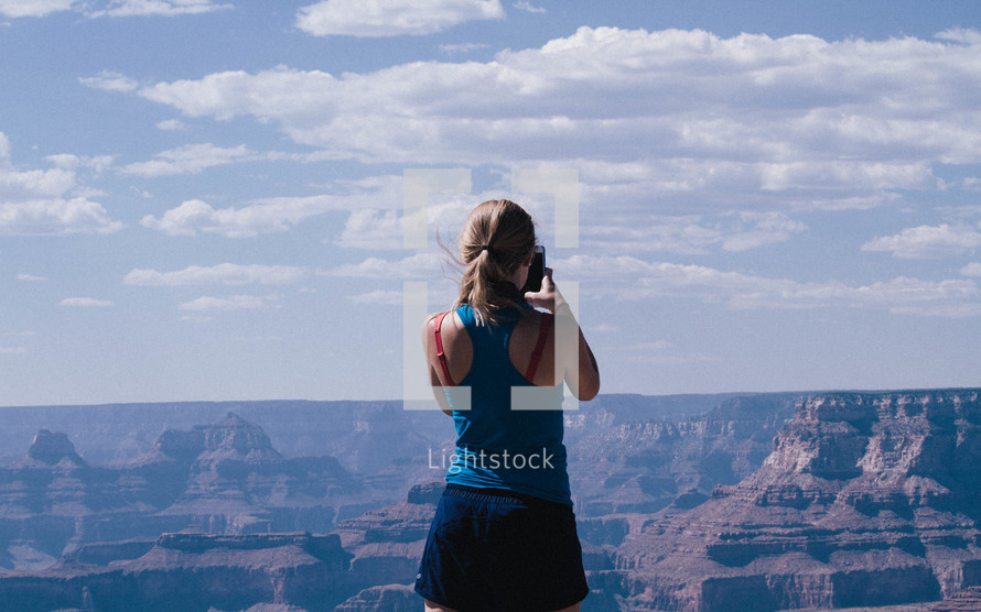 a woman standing at the edge of a cliff taking a picture with a cellphone camera 