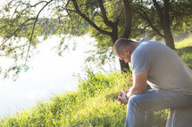 a man kneeling with his head bowed in prayer by a lake 