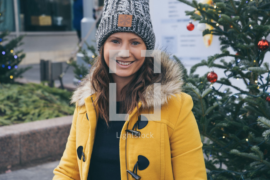 a young woman in a coat and beanie standing in front of a Christmas tree 