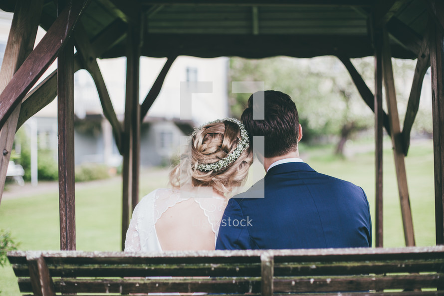 a bride and groom sitting on a bench outdoors with backs to the camera 