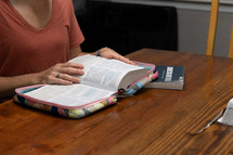 Woman in orange shirt sitting at kitchen table opening Bible next to study book during Bible study time in her discipleship group