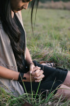 A young woman sitting in the grass with hands clasped and head bowed in prayer.