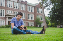 A man reads a Bible on a lawn outside an apartment building.