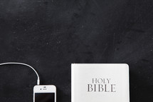 White Bible next to a white cellphone with ear buds plugged in on a chalkboard background.