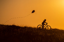 child riding a bike at sunset pulling a kite 