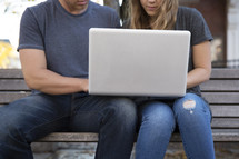 a couple sitting on a bench looking at a laptop computer screen 